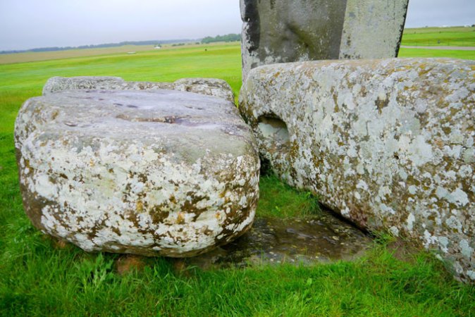 In this closeup photo, two mottled stones from Stonehenge lay on top of a darker brown stone that is embedded in the grass. That partially buried stone is the Altar Stone.